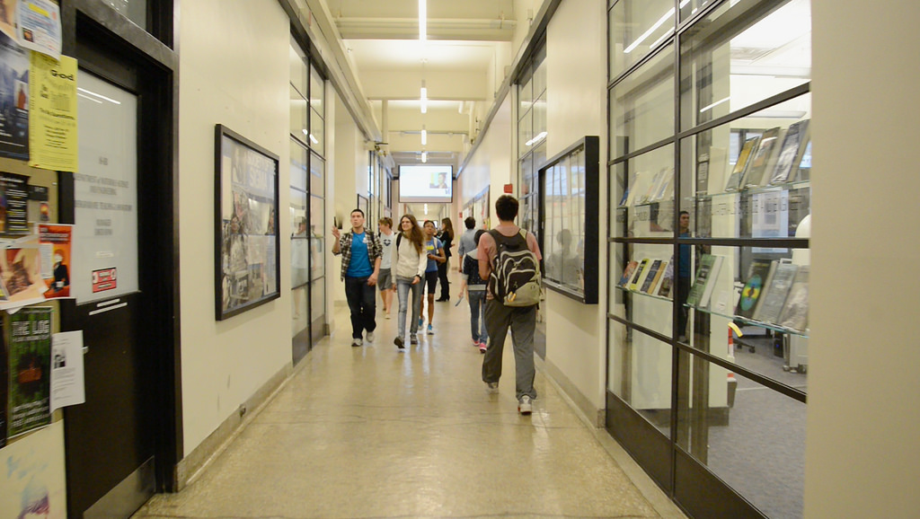 Students walk down the Infinite Corridor. There are windows into a lab on one side and posters on the opposite wall.