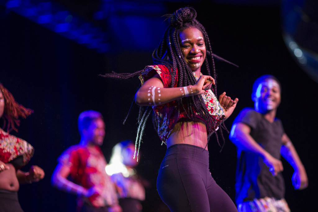 A performer dances in the foreground, with several more in the background. The stage is illuminated with blue light.