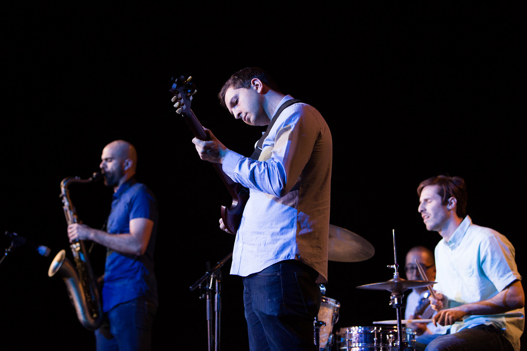 Three musicians play a saxophone, guitar, and the drums.