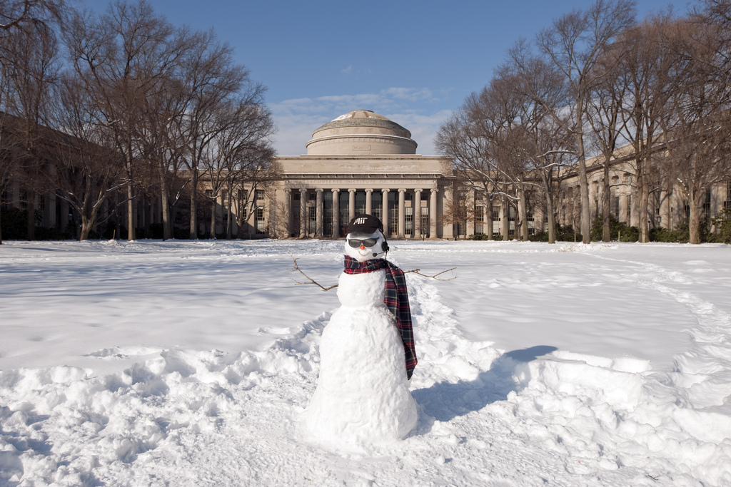 A snowman wearing a scarf, sunglasses, a headset, and an MIT hat stands in Killian Court, in front of the MIT Dome.