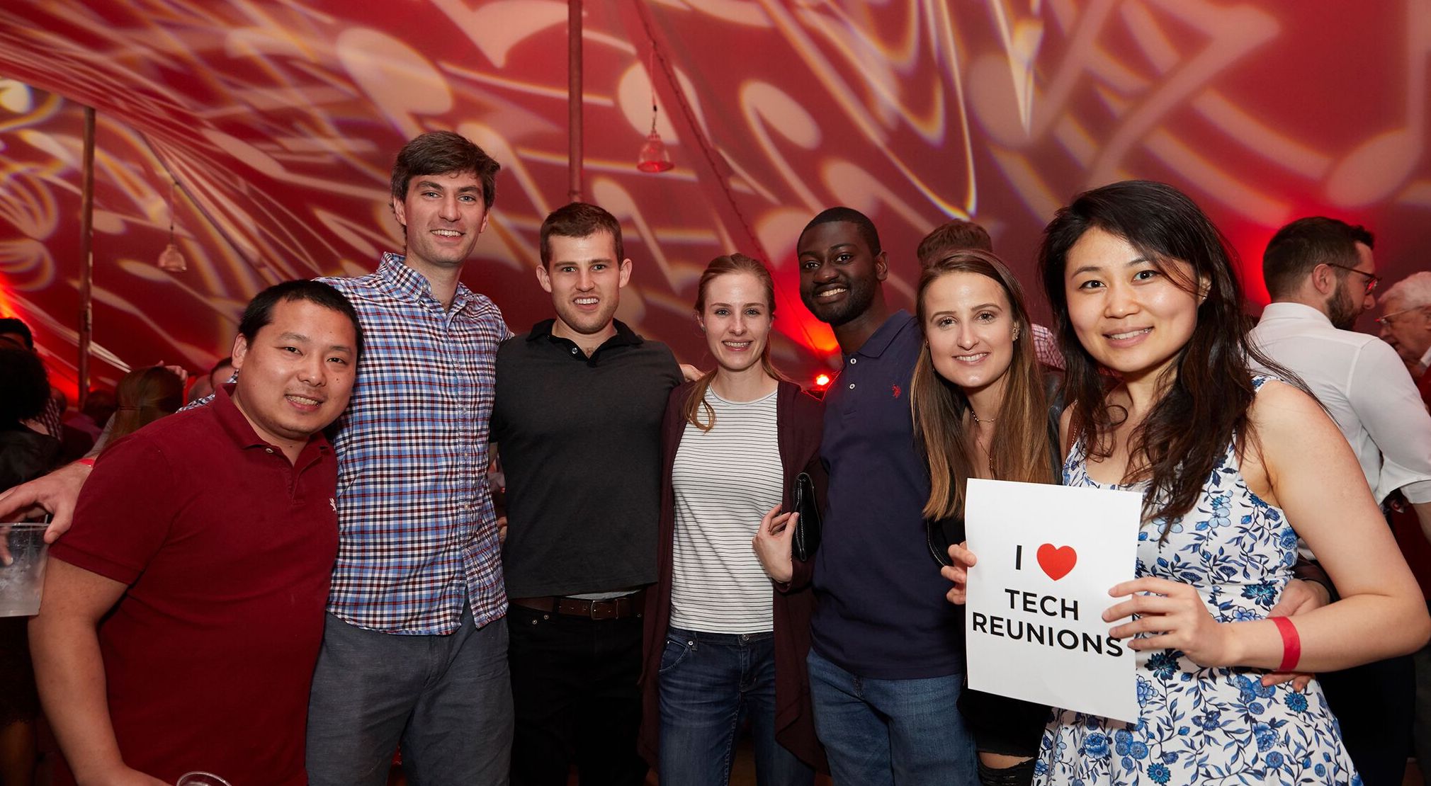 Seven alumni standing together. One person holds a sign that says, "I (heart) Tech Reunions."