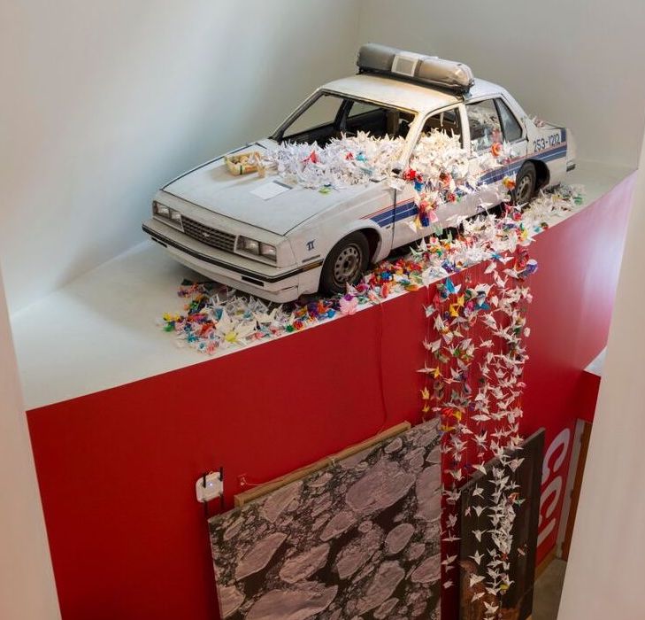 A police car with strands made up of colorful paper cranes coming out of the windows sits on a platform with a red wall.