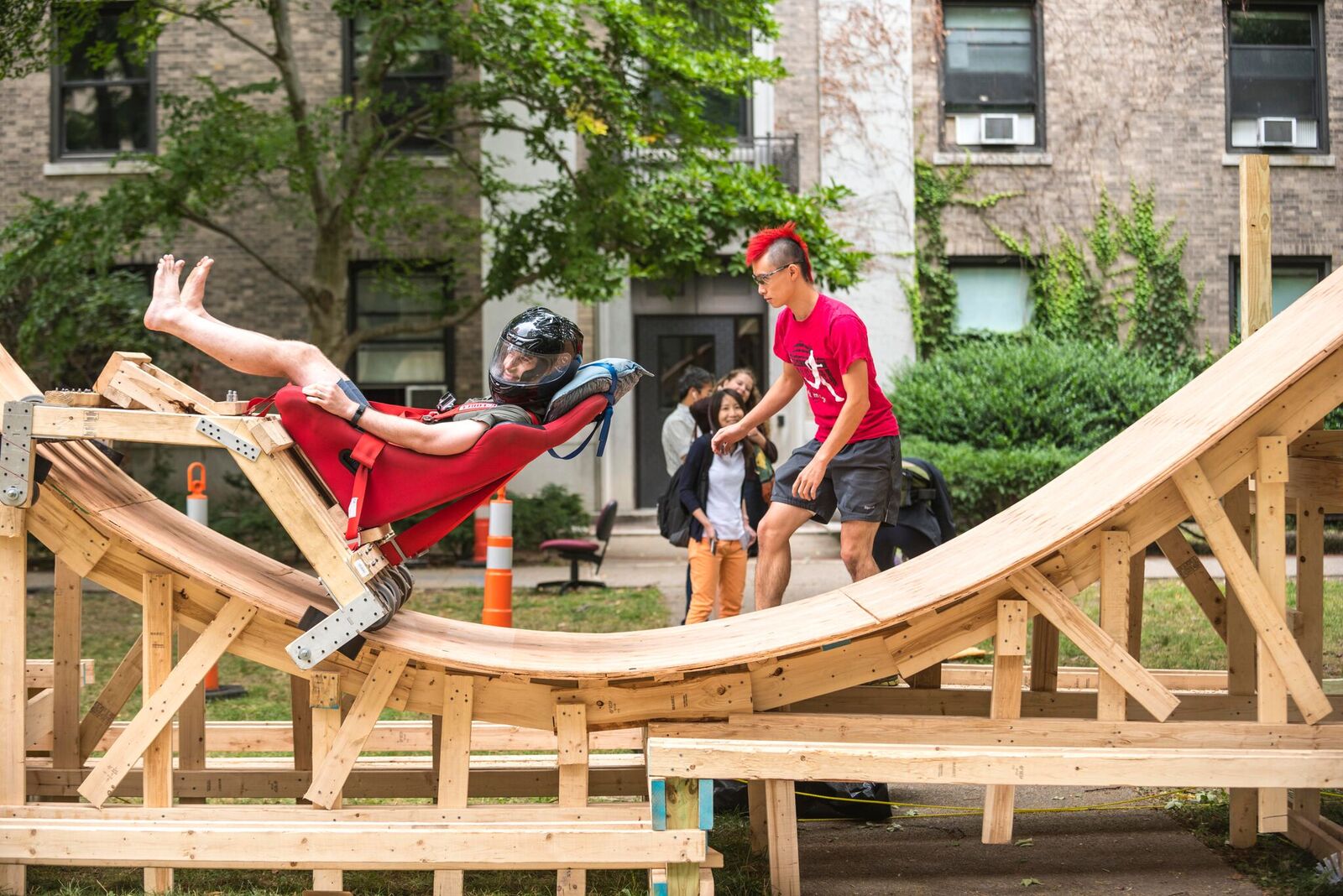 A student takes a ride in a wooden roller coaster while several other students look on. An MIT residence hall is in the background.