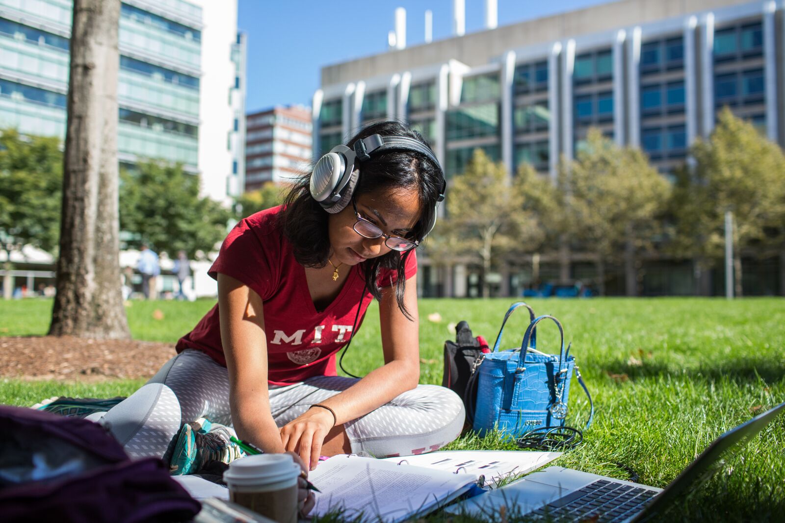A student wearing headphones and an MIT t-shirt sits on the grass while writing in a notebook. A few MIT buildings are in the background.