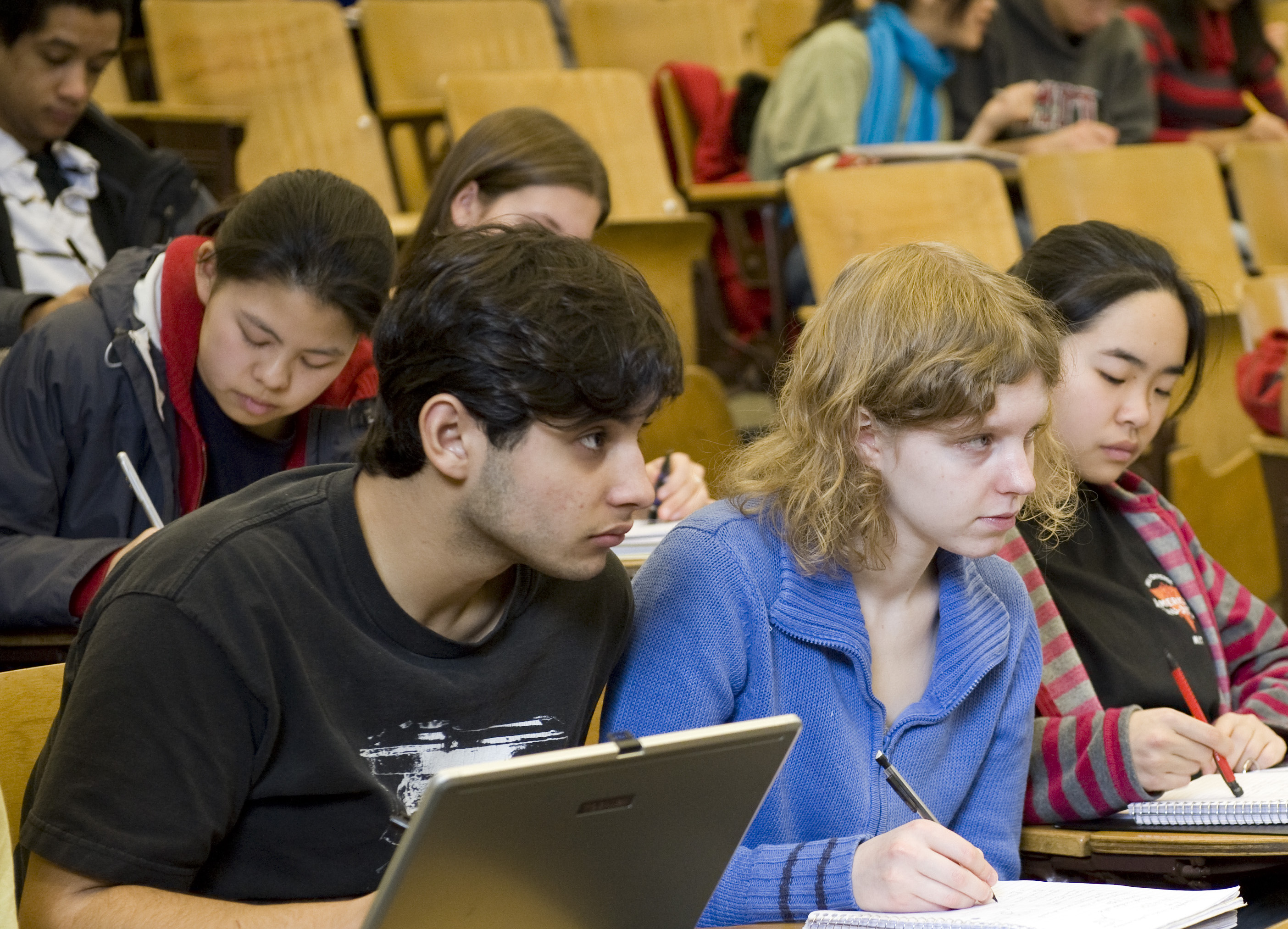 Students seated in a lecture hall with tiered seats. Two are looking at the front of the room and two other student are taking notes.