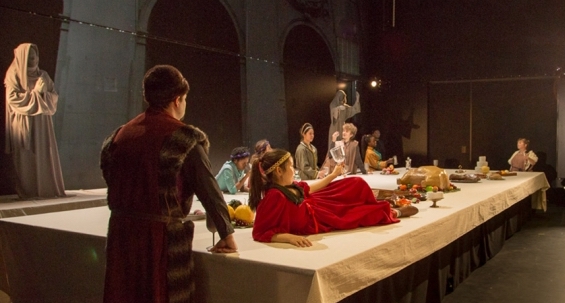 Several students on the set of a play. The student in front is wearing a long, red garment and lays on a table, holding a drinking glass.